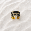 A wide open-band ring with a black and gold woven or beaded exterior and polished gold interior, photographed on a textured off-white surface.