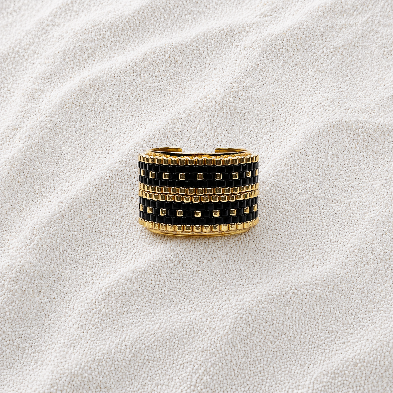 A wide gold-tone ring with three rows of black square stones, photographed on a textured white fabric background.
