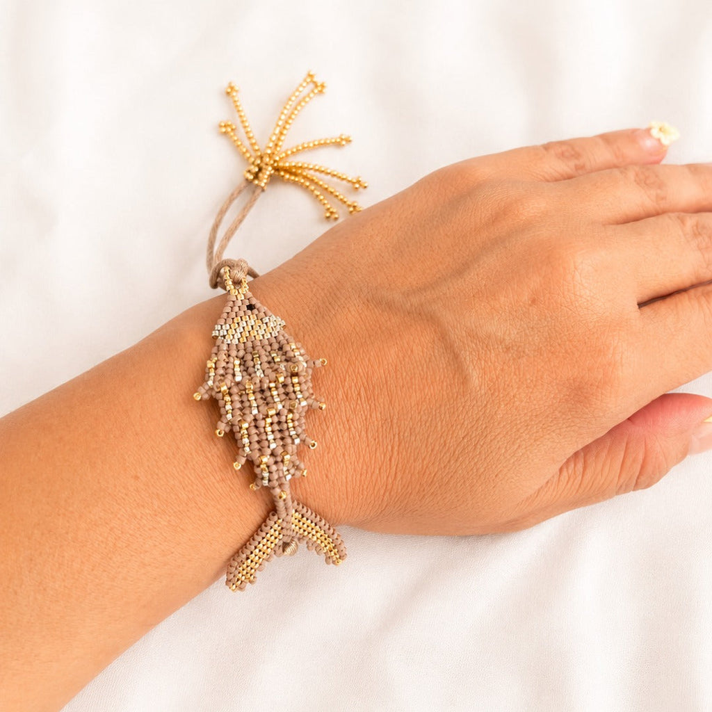 A gold beaded bracelet featuring a fish and starfish design, shown worn on a person's wrist against a white fabric background.