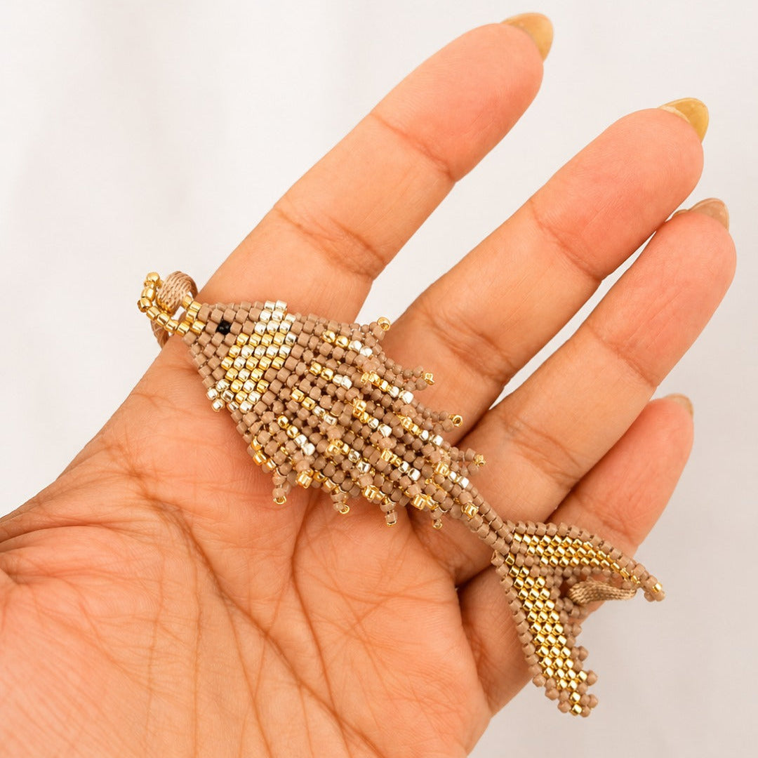 A hand holding a small fish-shaped beaded charm or brooch made of gold and beige seed beads, shown against a plain light background.