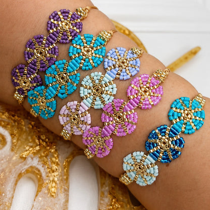 A close-up of a person's wrist wearing multiple beaded bracelets with gold-tone circular centers and rows of beads in purple, turquoise, light blue, pink, and dark blue. A white wall and gold decorative elements are softly blurred in the background.