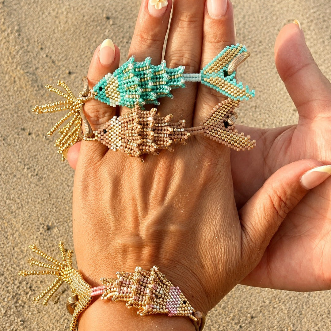 Three beaded fish-shaped bracelets in turquoise, gold, and beige are displayed on a person's hand against a sandy beach background.