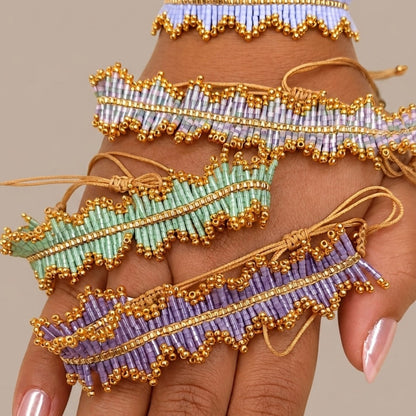 Three beaded finger bands with gold-tone borders and ties, shown wrapped around a hand. The bands are in pastel lavender, mint green, and light blue, set against a solid beige background.