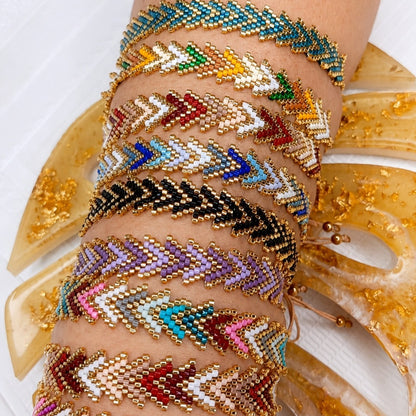 A stack of beaded chevron-patterned bracelets worn on a person's wrist, featuring gold-tone metal and multicolored seed beads in turquoise, green, yellow, red, blue, black, purple, pink, and white. The bracelets are styled against a background with translucent gold leaf monstera leaves on a white surface.