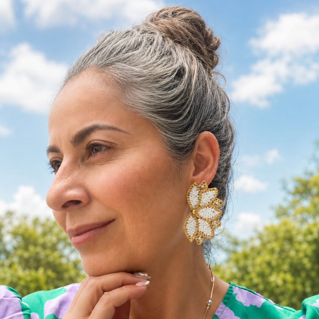 A woman with gray hair in a bun wearing a green and purple floral top and a gold flower-shaped earring, photographed outdoors against a blue sky with clouds.