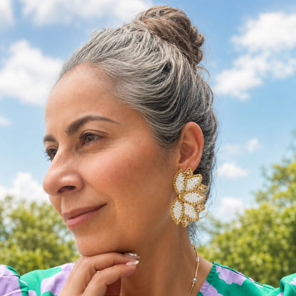 A woman with gray hair in a bun wearing a green and purple floral top and a gold flower-shaped earring, photographed outdoors against a blue sky with clouds.
