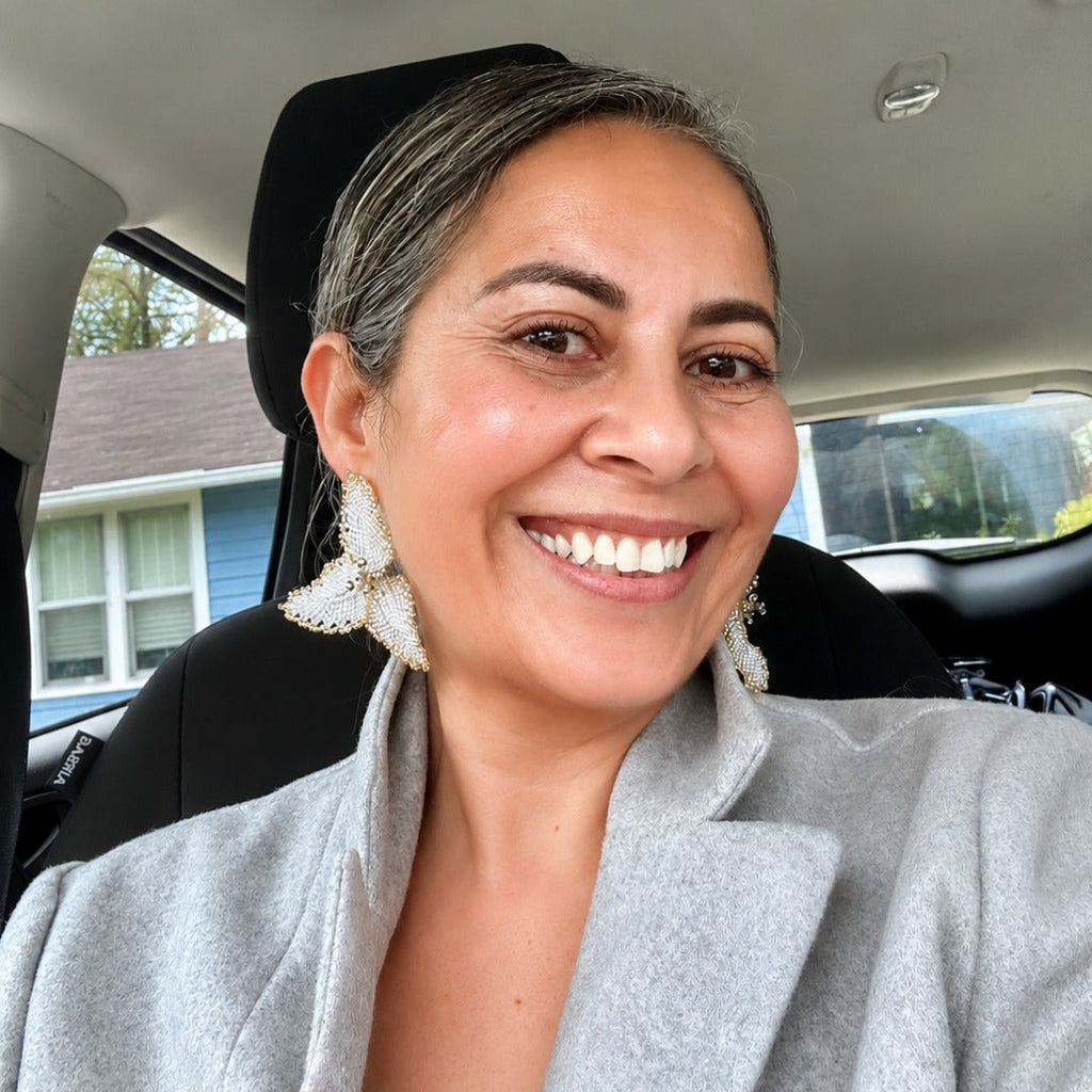A smiling woman with gray hair pulled back, wearing a light gray blazer and large white statement earrings, sitting inside a car.