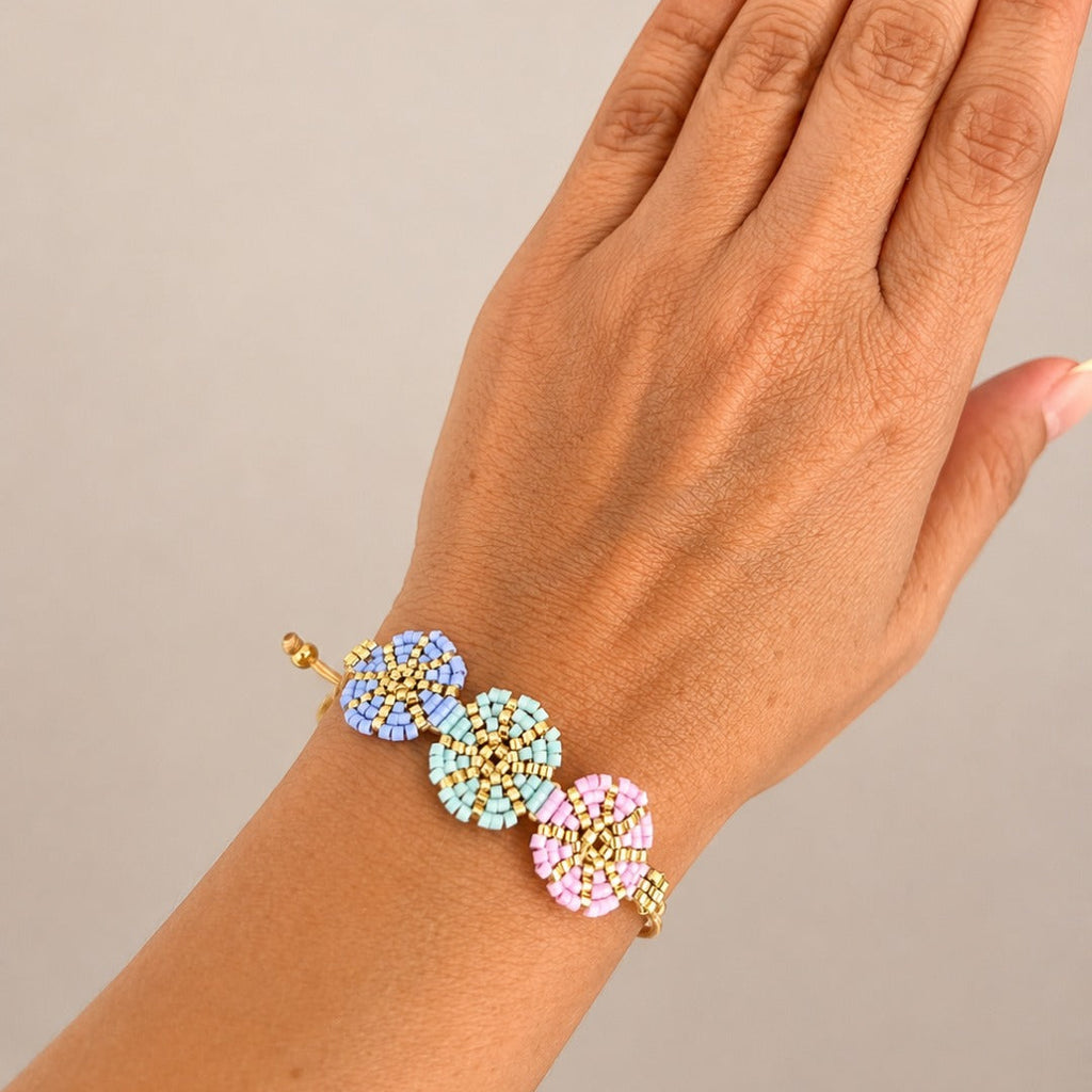 A close-up of a hand wearing a gold-tone beaded bracelet with three pastel flower clusters in blue, mint, and pink.