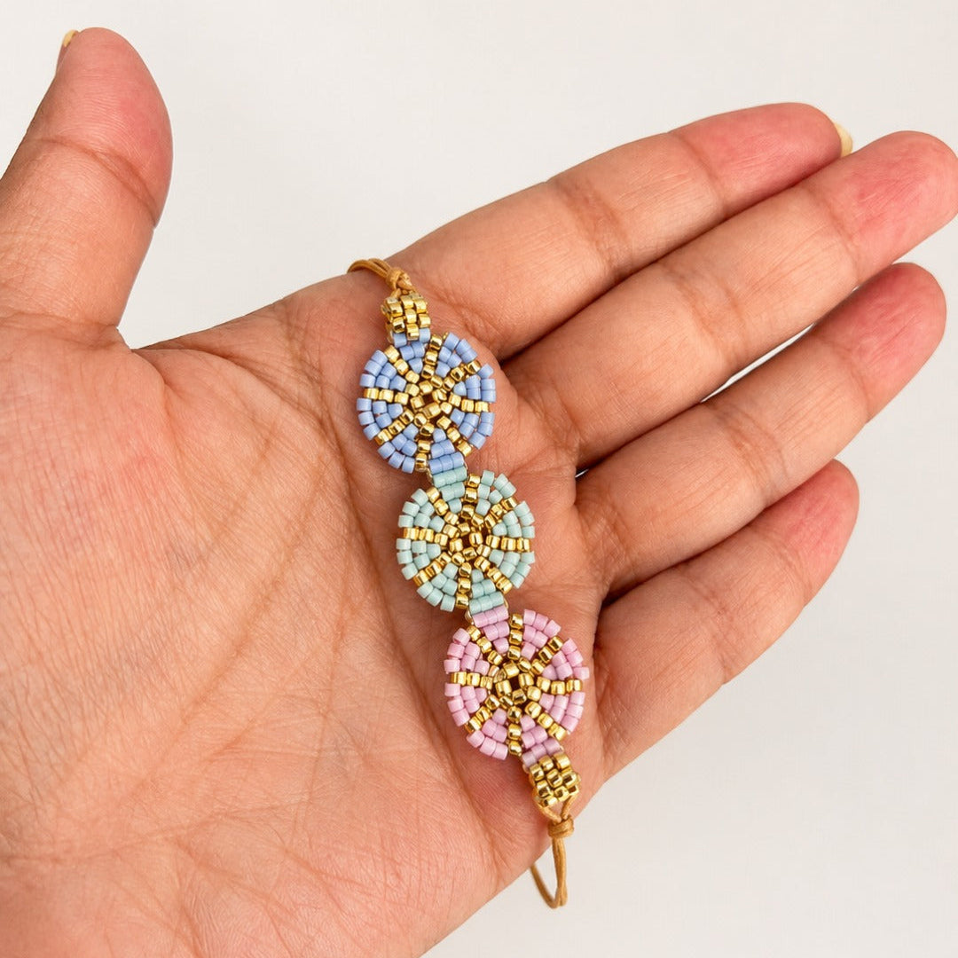 A handmade beaded bracelet with three circular floral clusters in pastel blue, light green, and pink, accented with gold seed beads and a gold wire loop, shown resting on a person's palm.