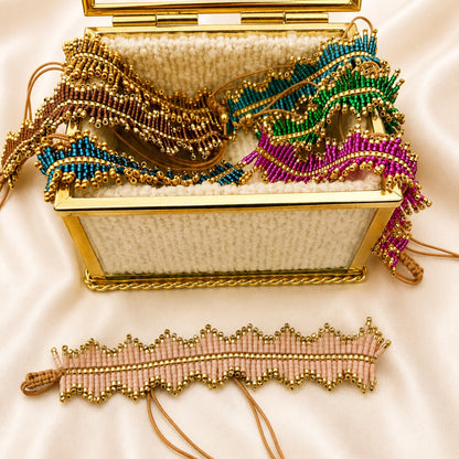 A gold-framed woven clutch bag with an open glass top, filled with colorful beaded fringe accessories, on a cream satin background. A matching pink and gold beaded wristlet lies in front.