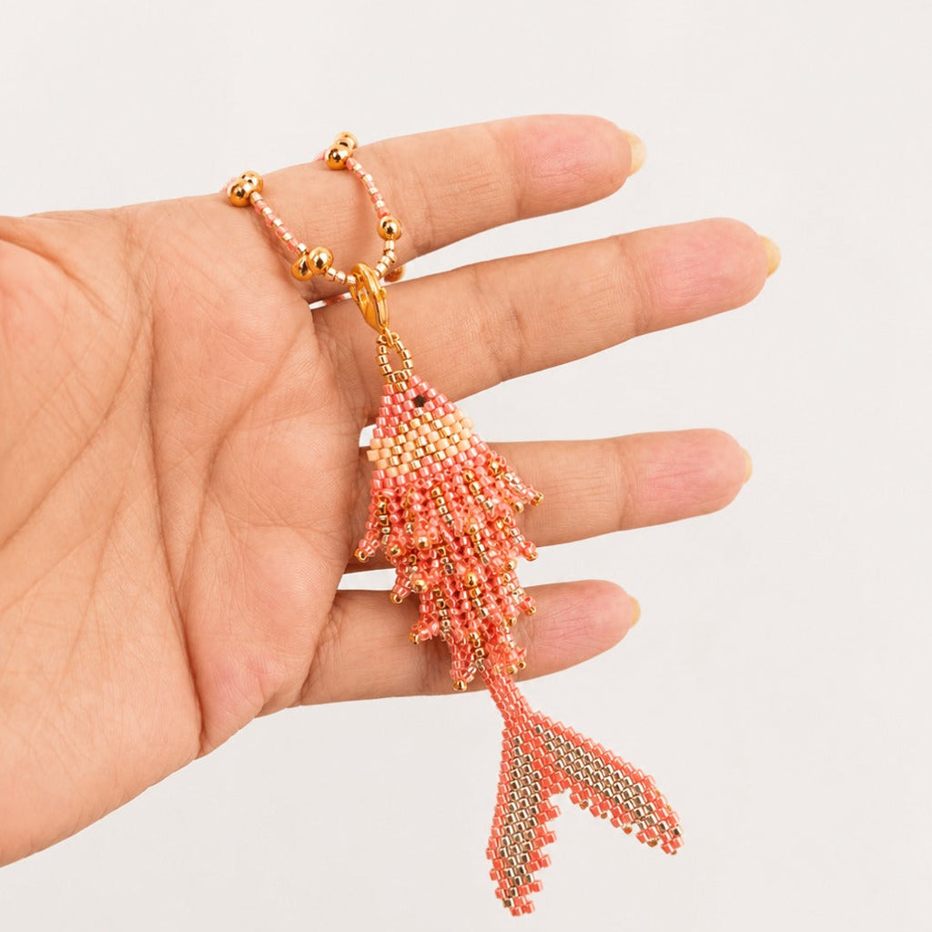 A coral pink and gold beaded fish-shaped pendant necklace held in a person's hand against a light gray background.