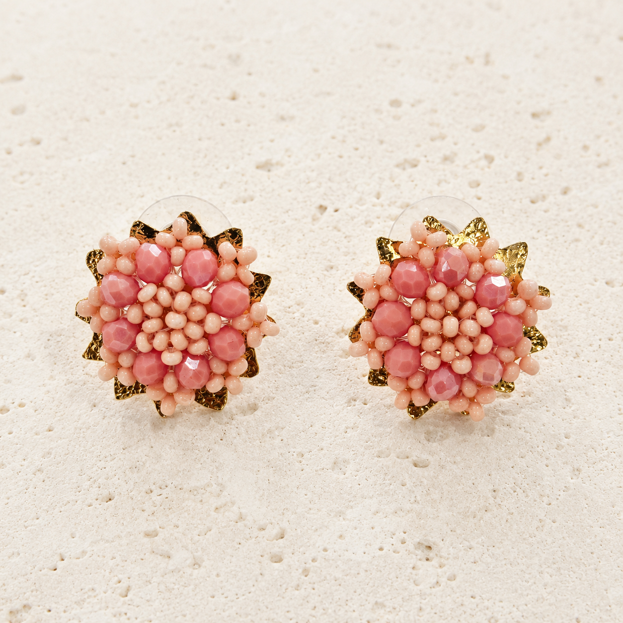 A pair of handmade beaded flower stud earrings with pink faceted beads, light pink seed beads, and gold glitter accents, displayed on a textured off-white surface.