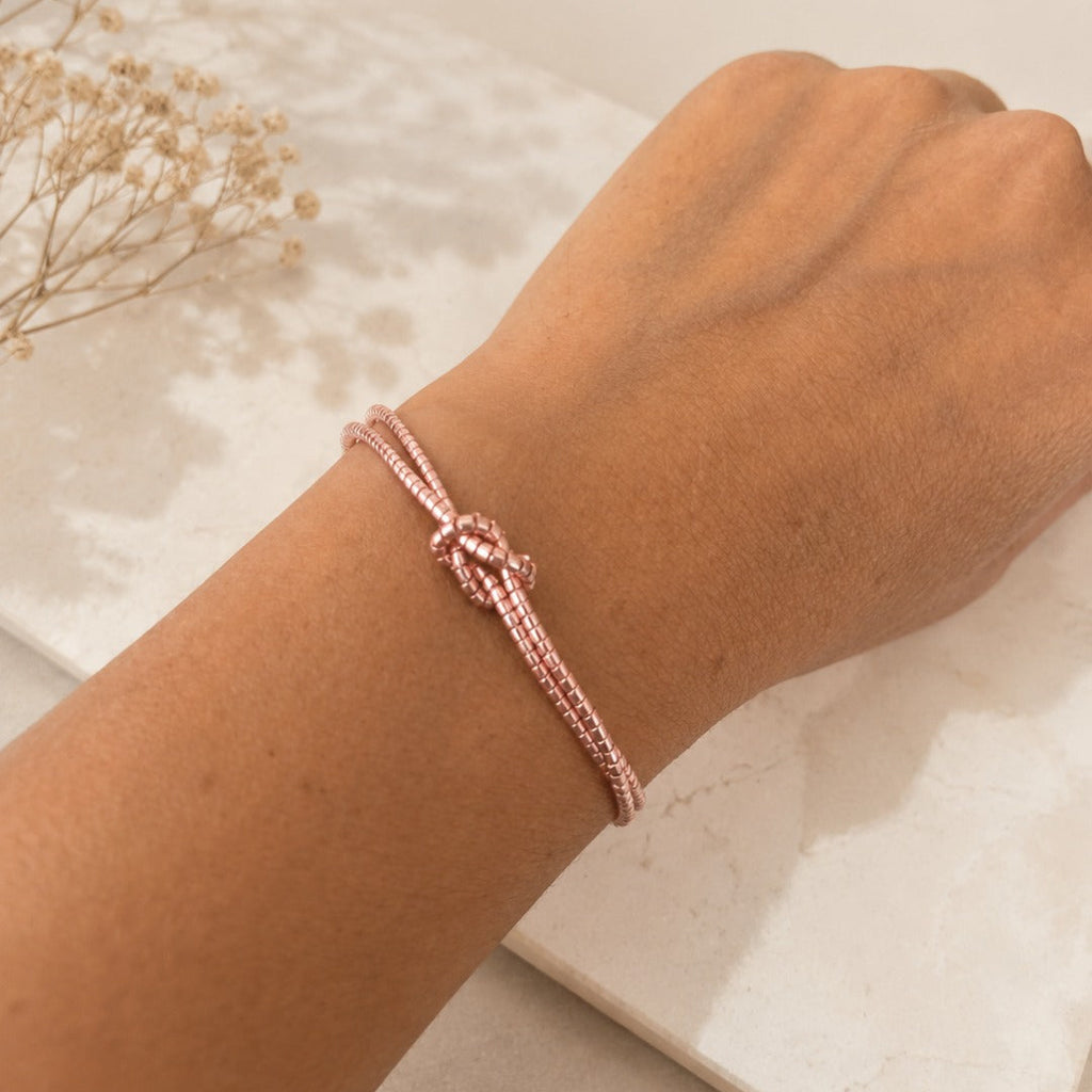A close-up of a person's wrist wearing a delicate rose gold braided cord bracelet with a knotted center detail, resting on a white marble slab with dried flowers in the background.