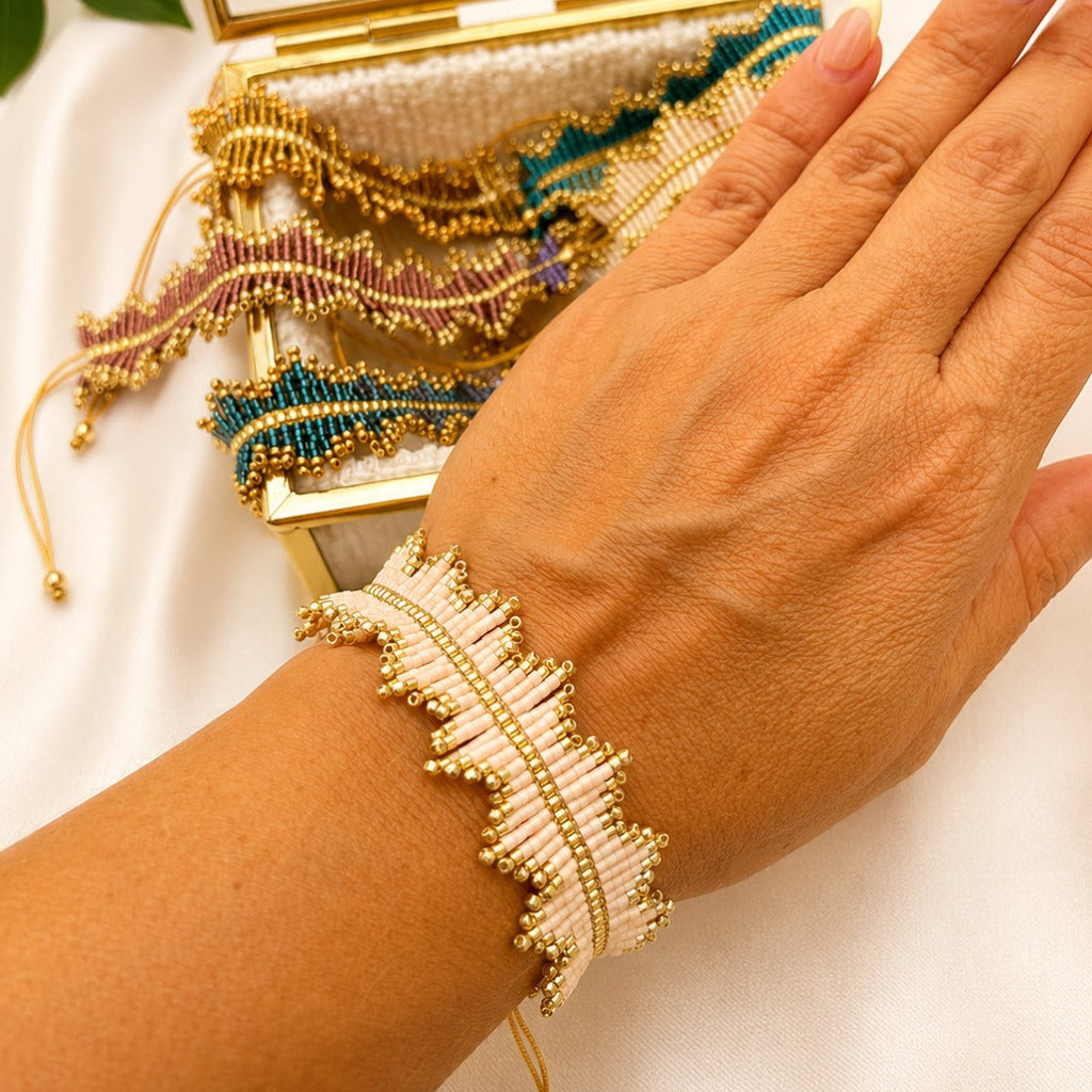 A close-up of a person's hand and wrist wearing a delicate beaded bracelet with cream beads and gold seed beads. An open gold jewelry box with more colorful beaded bracelets sits behind the hand on a white fabric background.