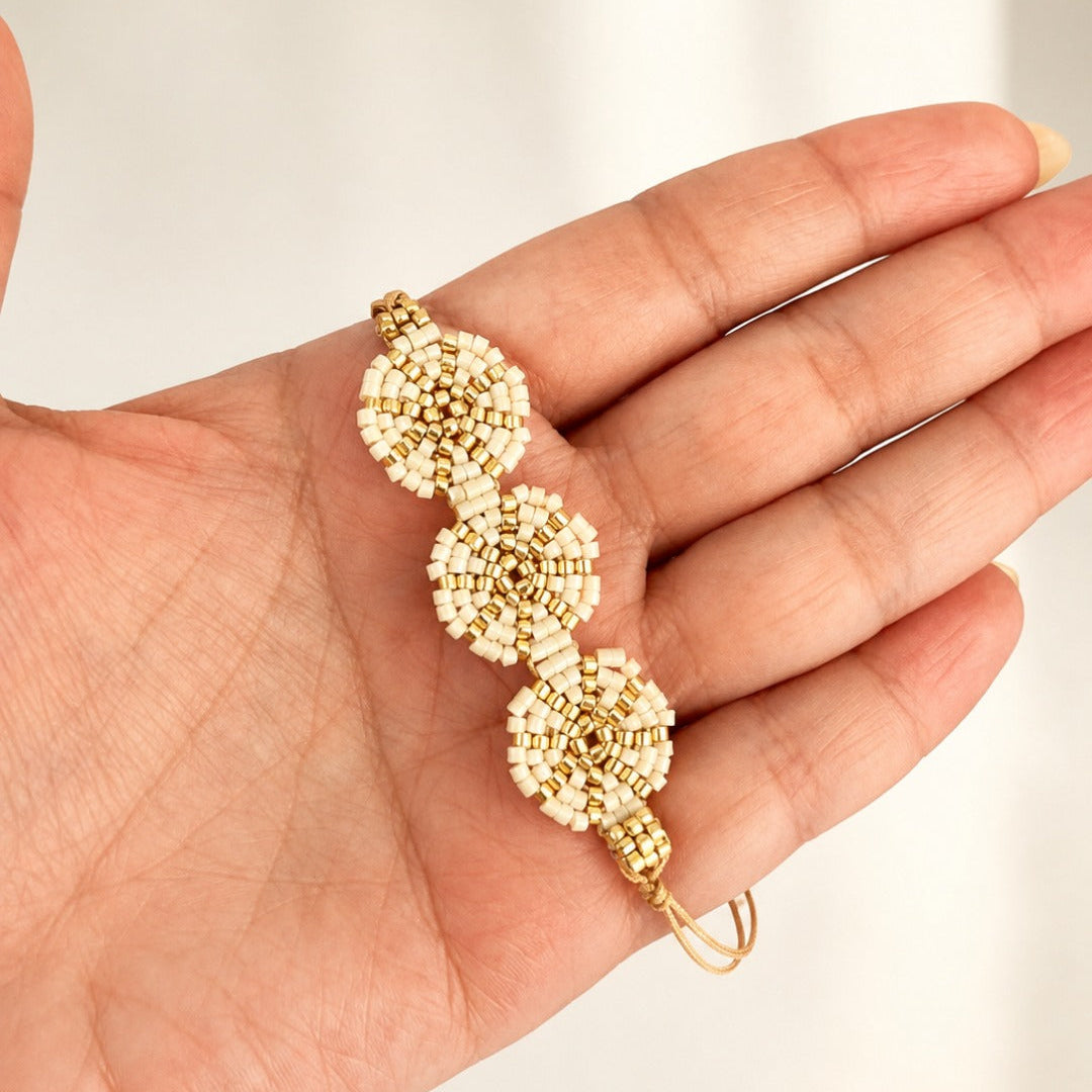A gold beaded bracelet with three circular floral clusters and a small loop at the end, held in a person's hand against a soft light background.