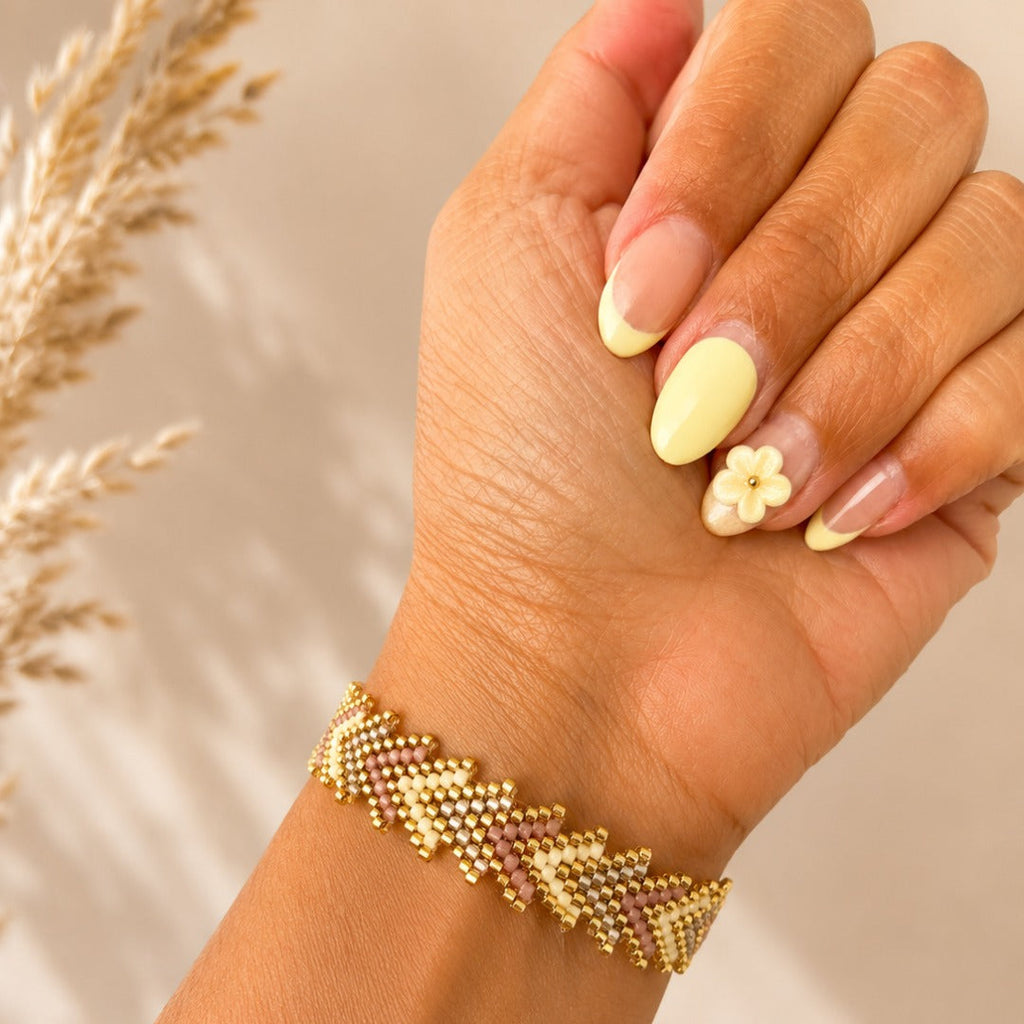 A close-up of a hand wearing a gold beaded chevron-patterned bracelet, with manicured nails and a small flower nail art accent, against a warm neutral background with dried grass.