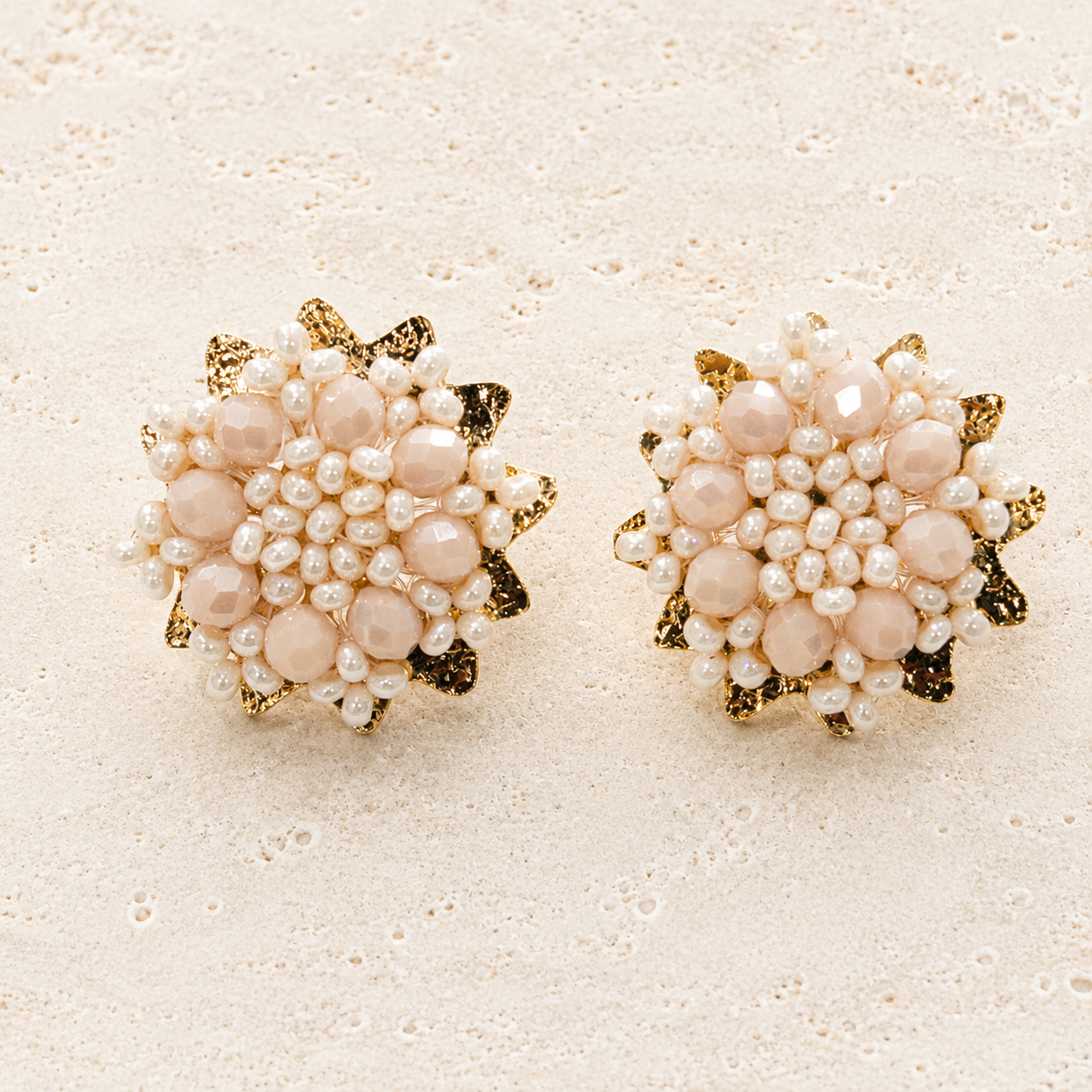A pair of round floral stud earrings featuring pale pink faux pearls, small white pearls, and gold-tone metal accents, displayed on a textured beige surface.