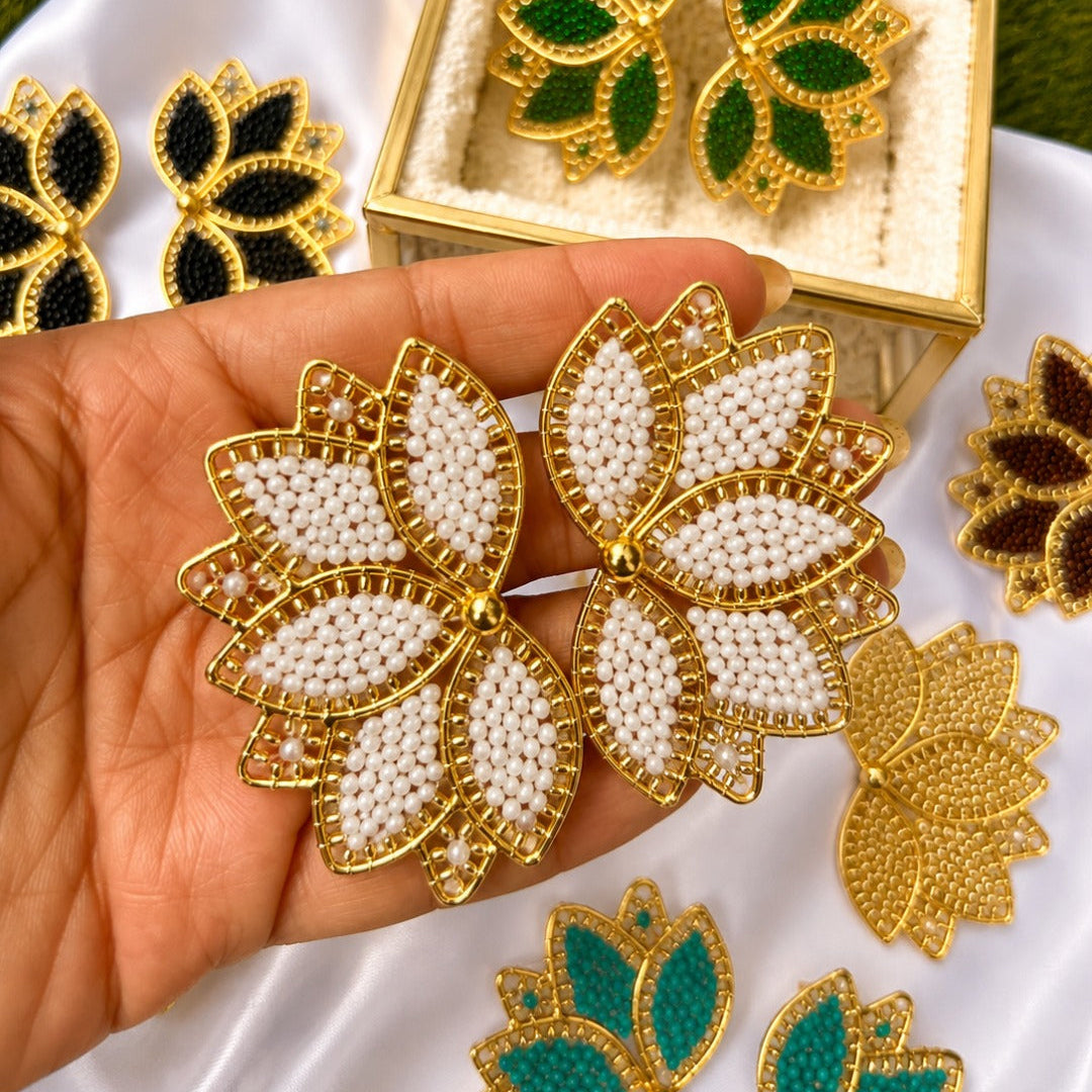 Gold-toned leaf-shaped earrings with white beadwork and green stone accents, shown in a hand with additional matching earrings and a display box on a white cloth.