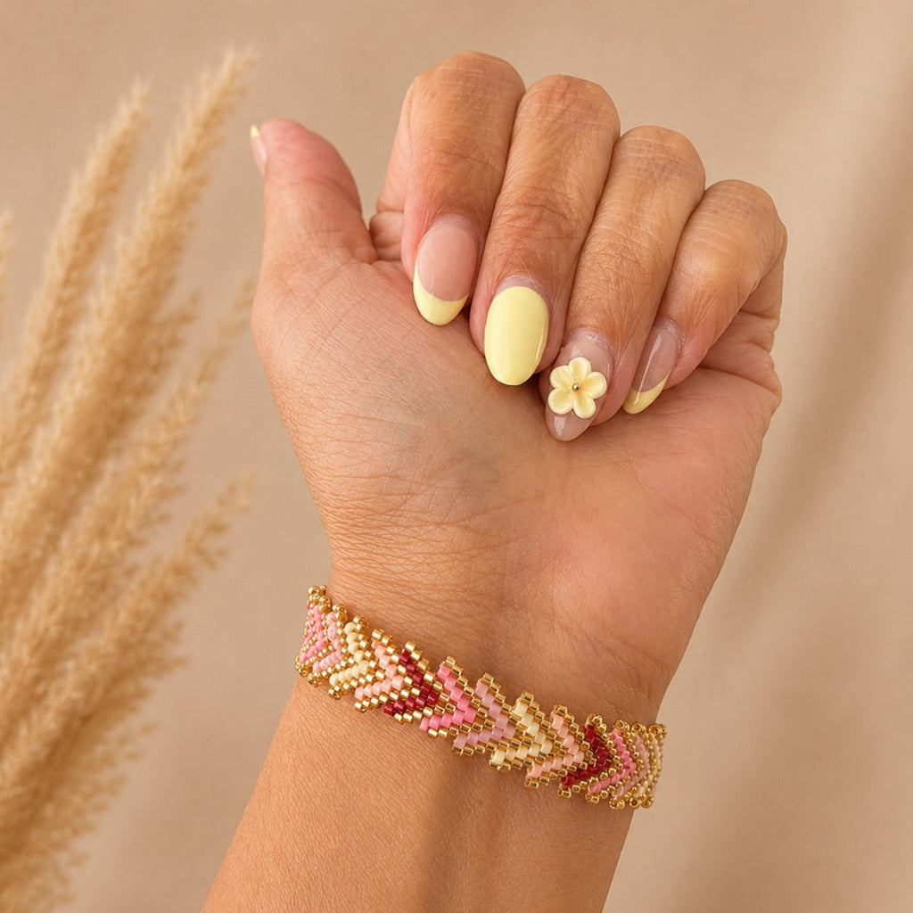 A close-up of a person's hand and wrist wearing a beaded chevron bracelet in gold, pink, and red tones, with neutral-toned dried grass stems in the background.