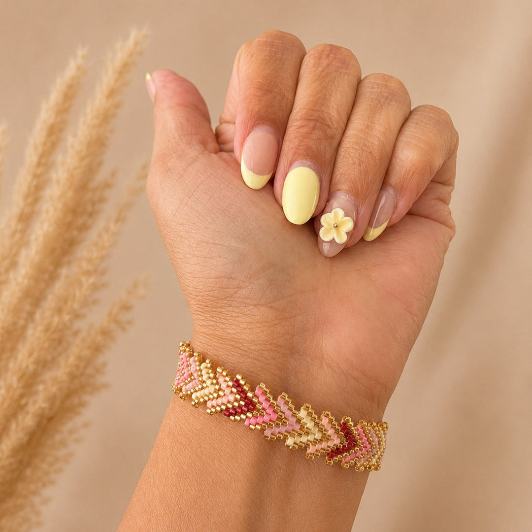 A close-up of a person's hand and wrist wearing a beaded chevron bracelet in gold, pink, and red tones, with neutral-toned dried grass stems in the background.