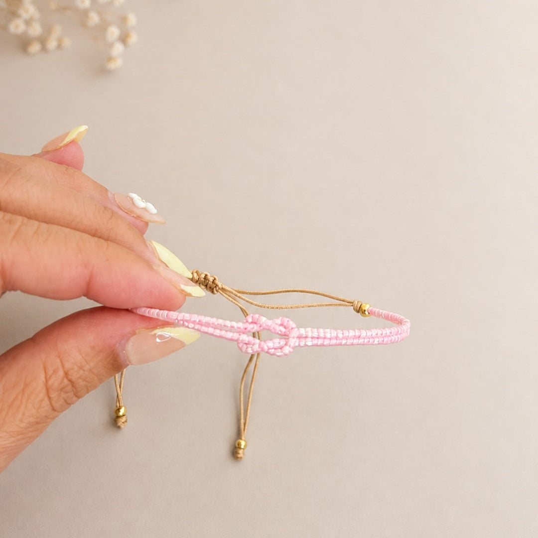 A hand with pale yellow manicured nails holds a delicate pink beaded bracelet with gold-toned adjustable cords and beads, set against a warm beige background with dried flowers in the upper left corner.