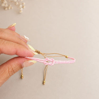 A hand with pale yellow manicured nails holds a delicate pink beaded bracelet with gold-toned adjustable cords and beads, set against a warm beige background with dried flowers in the upper left corner.