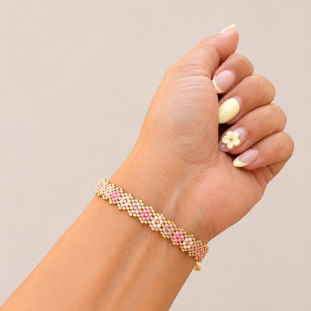 A gold chain bracelet with alternating pink and white floral links, shown worn on a person's wrist against a solid beige background.