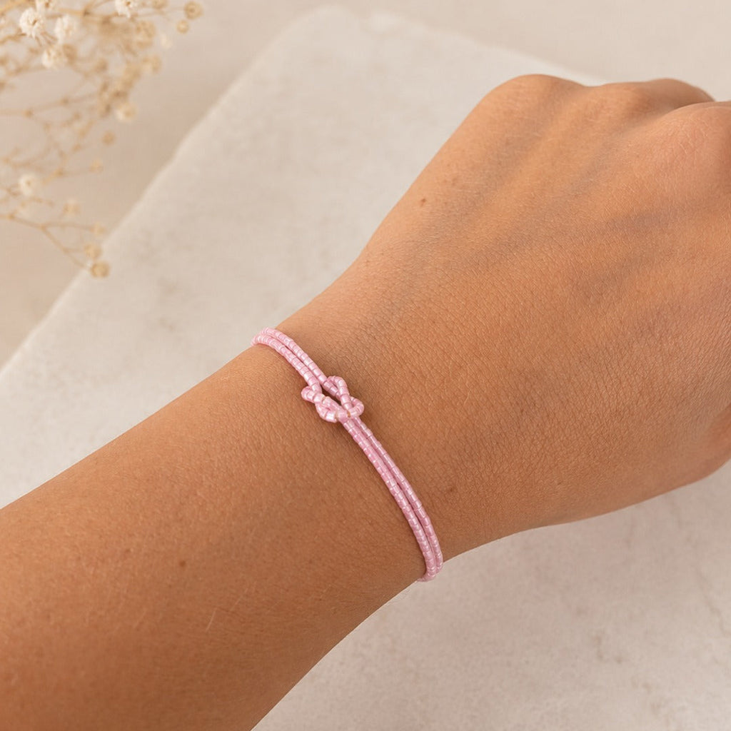 A delicate light pink braided cord bracelet with a small bow detail, shown worn on a person's wrist against a soft light beige background.