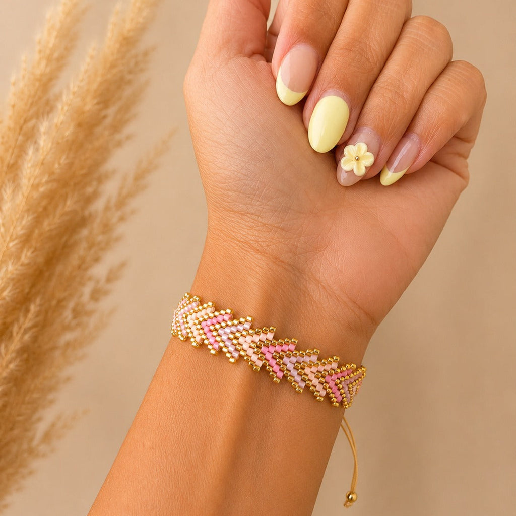A close-up of a hand wearing a delicate beaded bracelet with pink, yellow, and gold beads, styled with a neutral beige background and dried pampas grass.