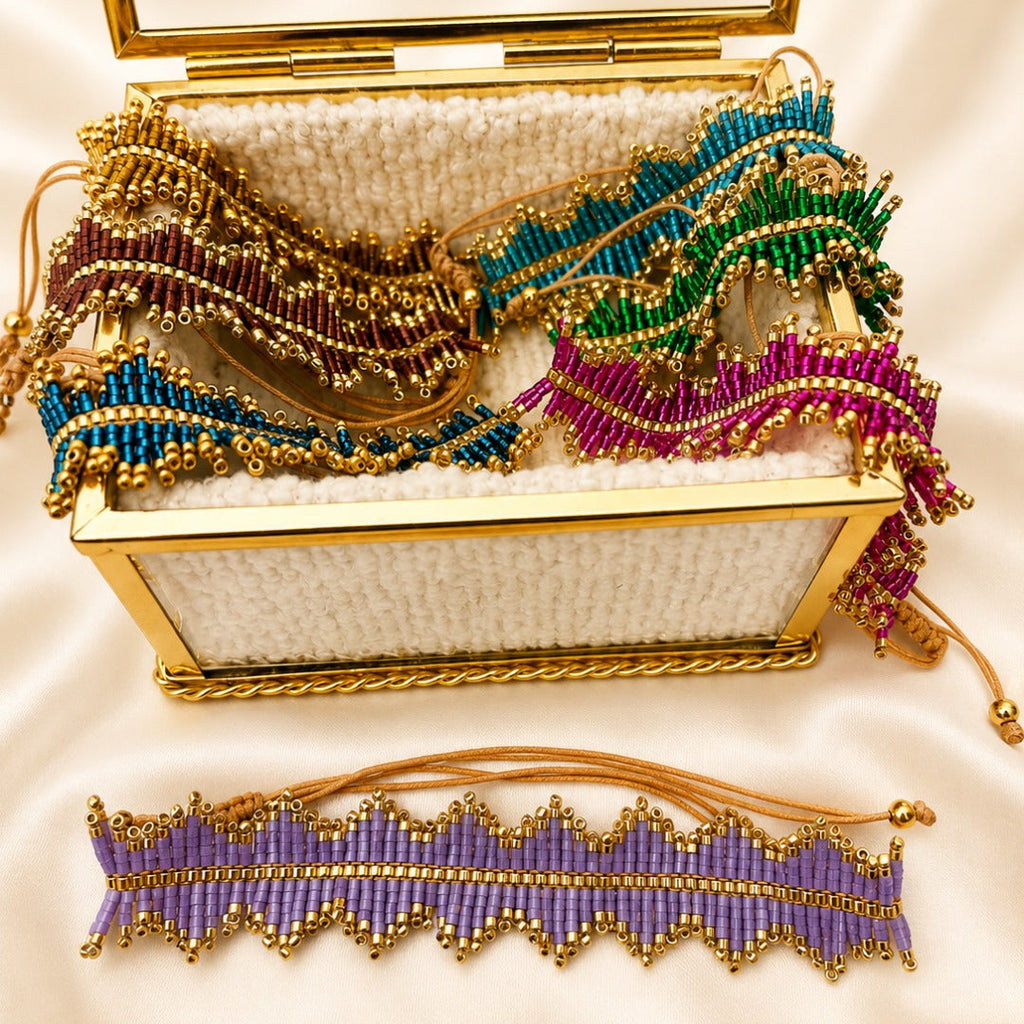 A gold-trimmed open jewelry box filled with colorful beaded accessories, including a purple beaded belt or anklet displayed in front on a cream satin background.