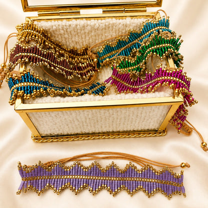 A gold-trimmed open jewelry box filled with colorful beaded accessories, including a purple beaded belt or anklet displayed in front on a cream satin background.