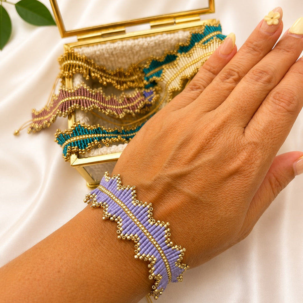 A close-up of a person's hand wearing a lavender and gold beaded leaf bracelet, with a matching gold clutch bag in the background on a white satin surface.