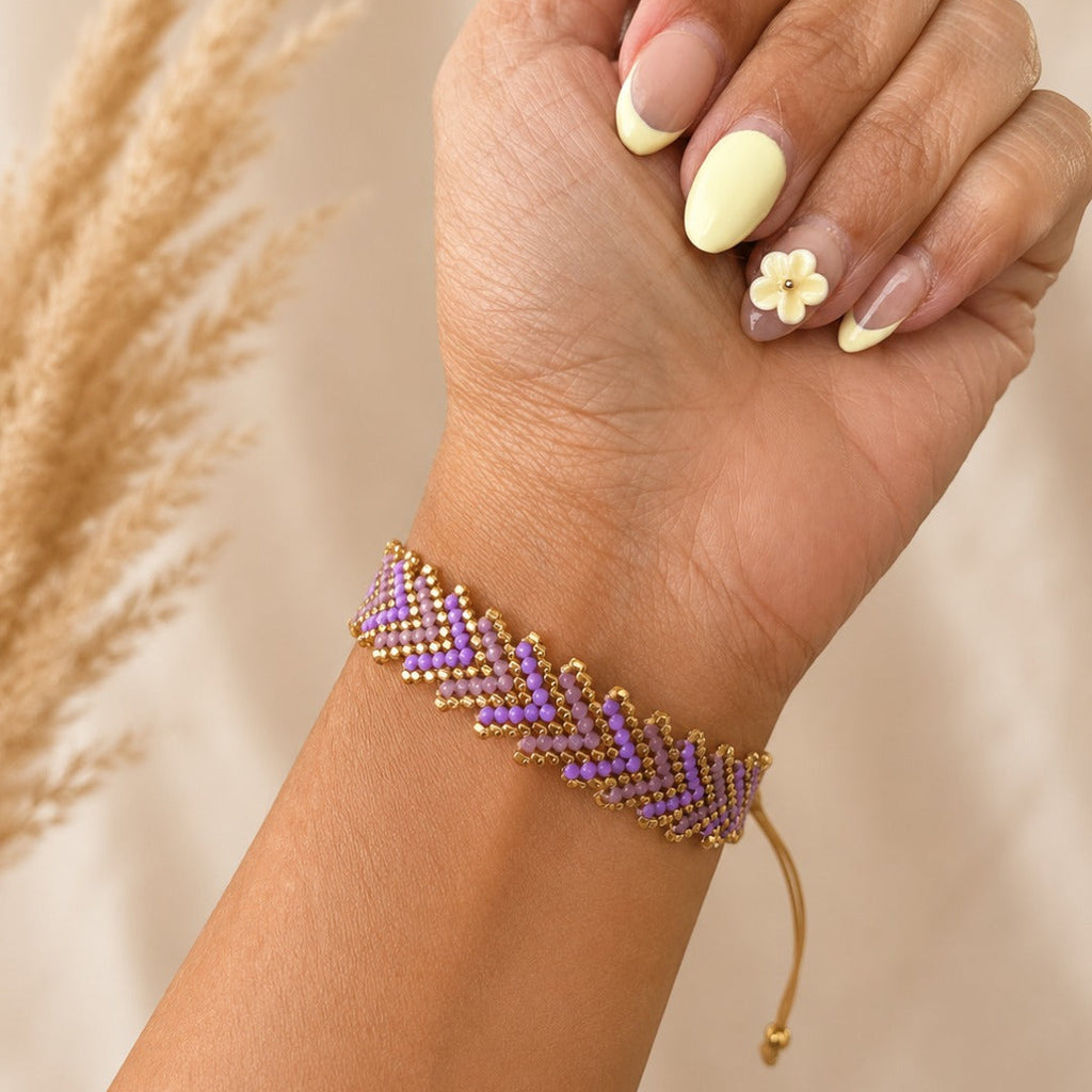 A close-up of a person's hand and wrist wearing a beaded chevron bracelet in lavender purple and gold. The hand has pale yellow manicured nails with a small flower detail on one nail, set against a soft beige background with blurred pampas grass on the left.