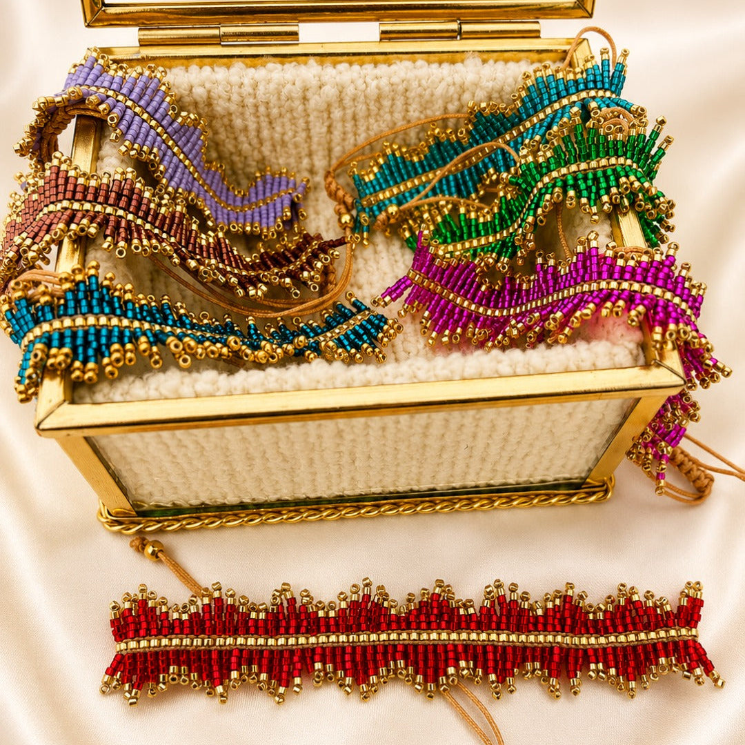 A gold-framed open jewelry box filled with colorful beaded accessories, including a purple and teal beaded headband, a green and teal beaded headband, a magenta and teal beaded headband, and a red and gold beaded wristlet bracelet, all arranged on a cream satin background.