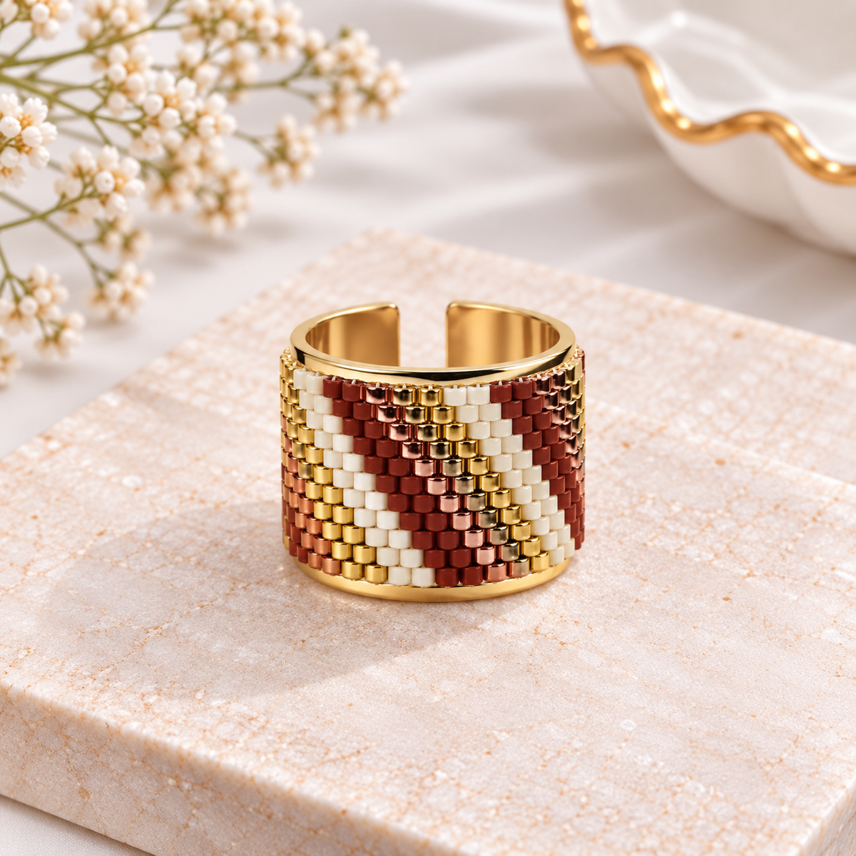 A wide open cuff ring with a woven geometric pattern in gold, rust red, and cream, displayed on a textured beige stone slab with baby's breath flowers in the background.