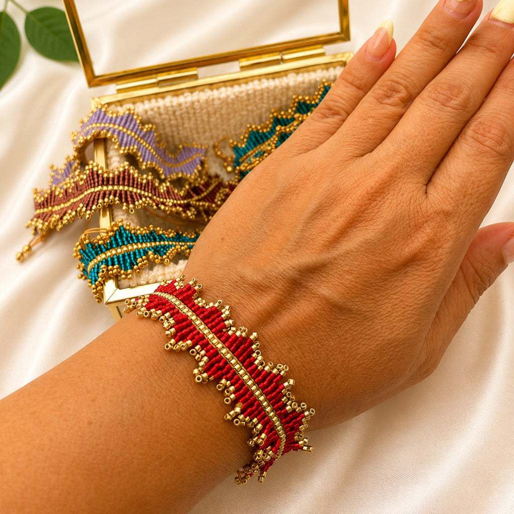 A close-up of a person's hand wearing a red and gold beaded bracelet, with additional colorful beaded bracelets and a gold-framed clutch resting on a white satin background.