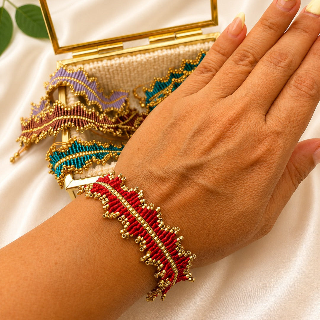 A close-up of a person's hand wearing a red and gold beaded bracelet, with additional colorful beaded bracelets and a gold-framed clutch resting on a white satin background.