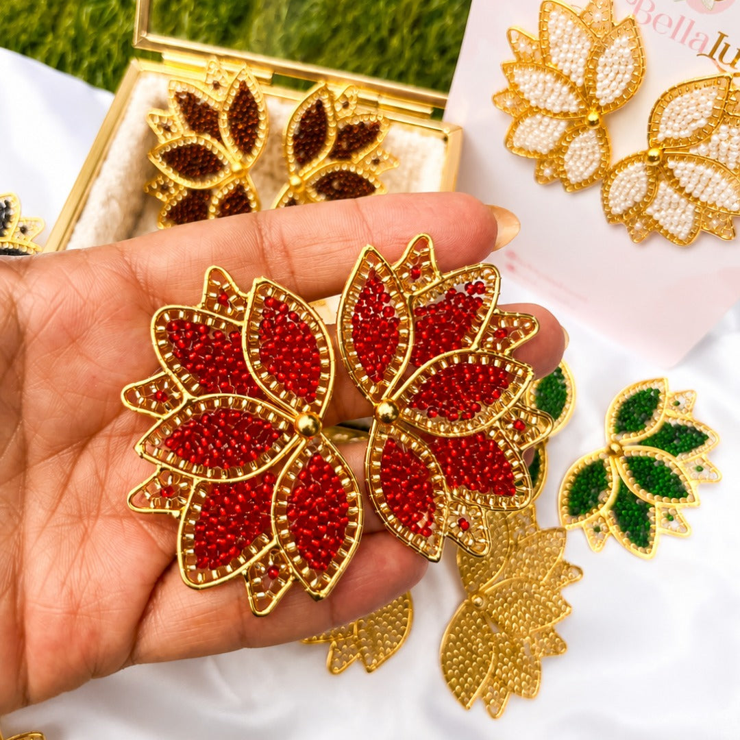 A hand holds a pair of gold-tone floral stud earrings embellished with red rhinestones, with additional multicolor floral earrings displayed on white fabric and a card in the background.