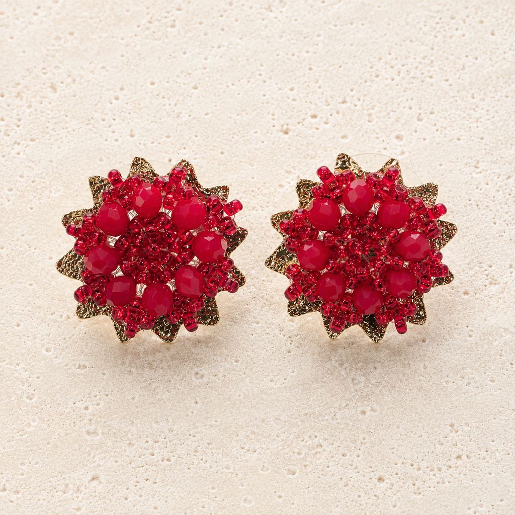 A pair of round cluster stud earrings featuring vibrant red faceted gemstones arranged in a floral or sunburst pattern with textured gold-toned edges, displayed on a light beige textured surface.
