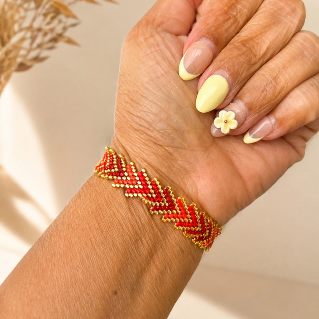 A close-up of a hand wearing a red and gold beaded chevron bracelet, with manicured nails and a small flower nail art accent.