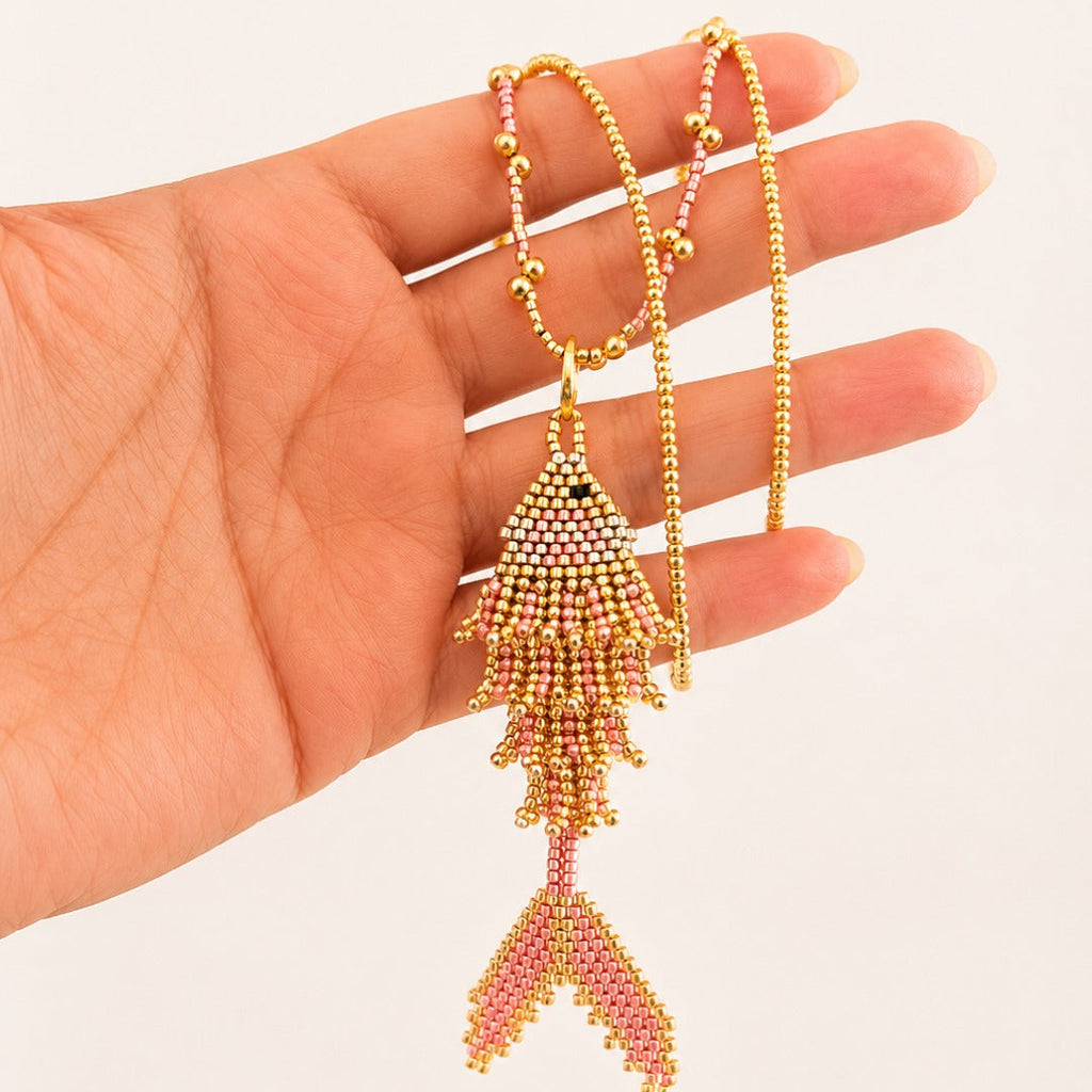 A hand holding a gold beaded necklace with a fish-shaped pendant and a dangling pink beaded tail against a plain light background.