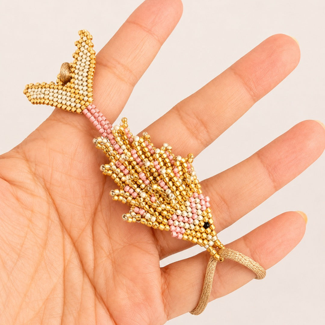 A hand holding a small beaded fish-shaped accessory in gold, pink, and white seed beads against a plain light background.
