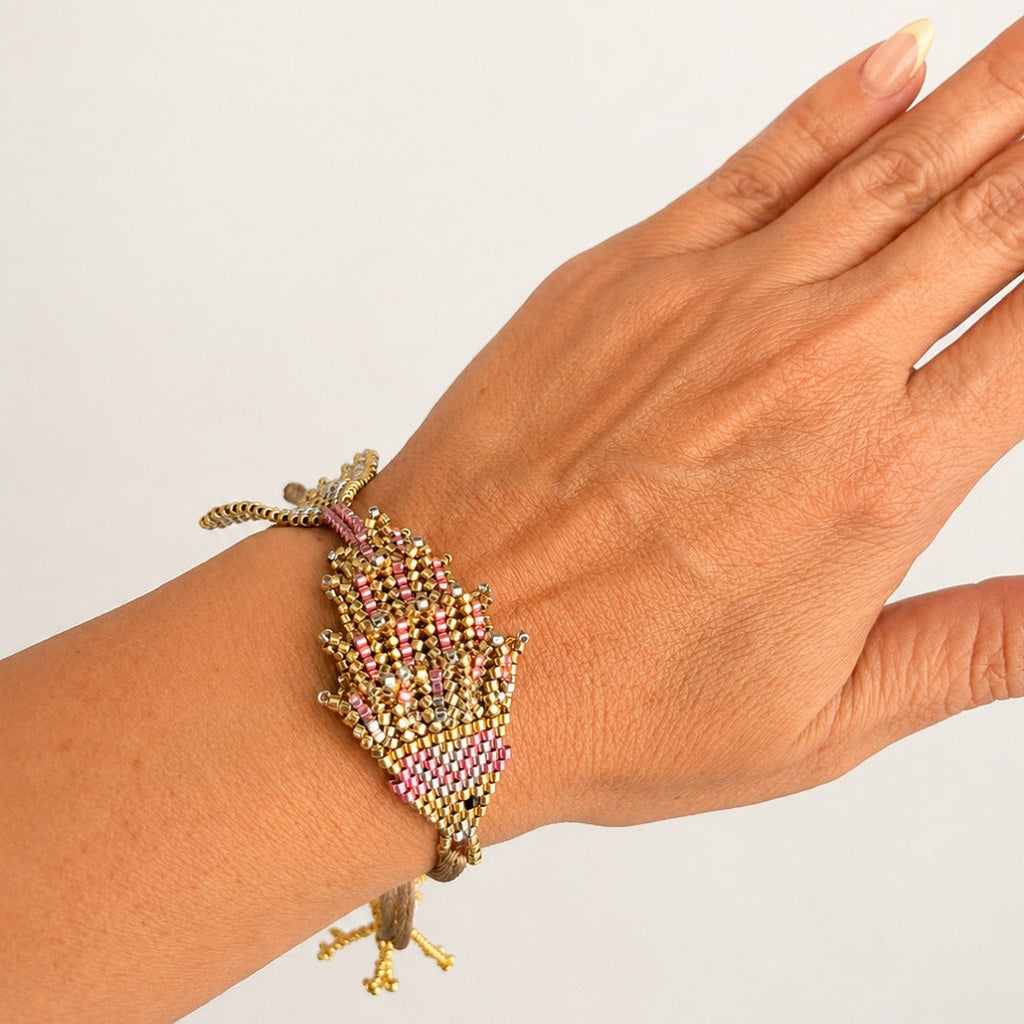 A close-up of a hand wearing a detailed beaded leaf-shaped bracelet with gold-tone metal and pink, purple, and green seed beads.