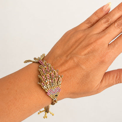 A close-up of a hand wearing a detailed beaded leaf-shaped bracelet with gold-tone metal and pink, purple, and green seed beads.