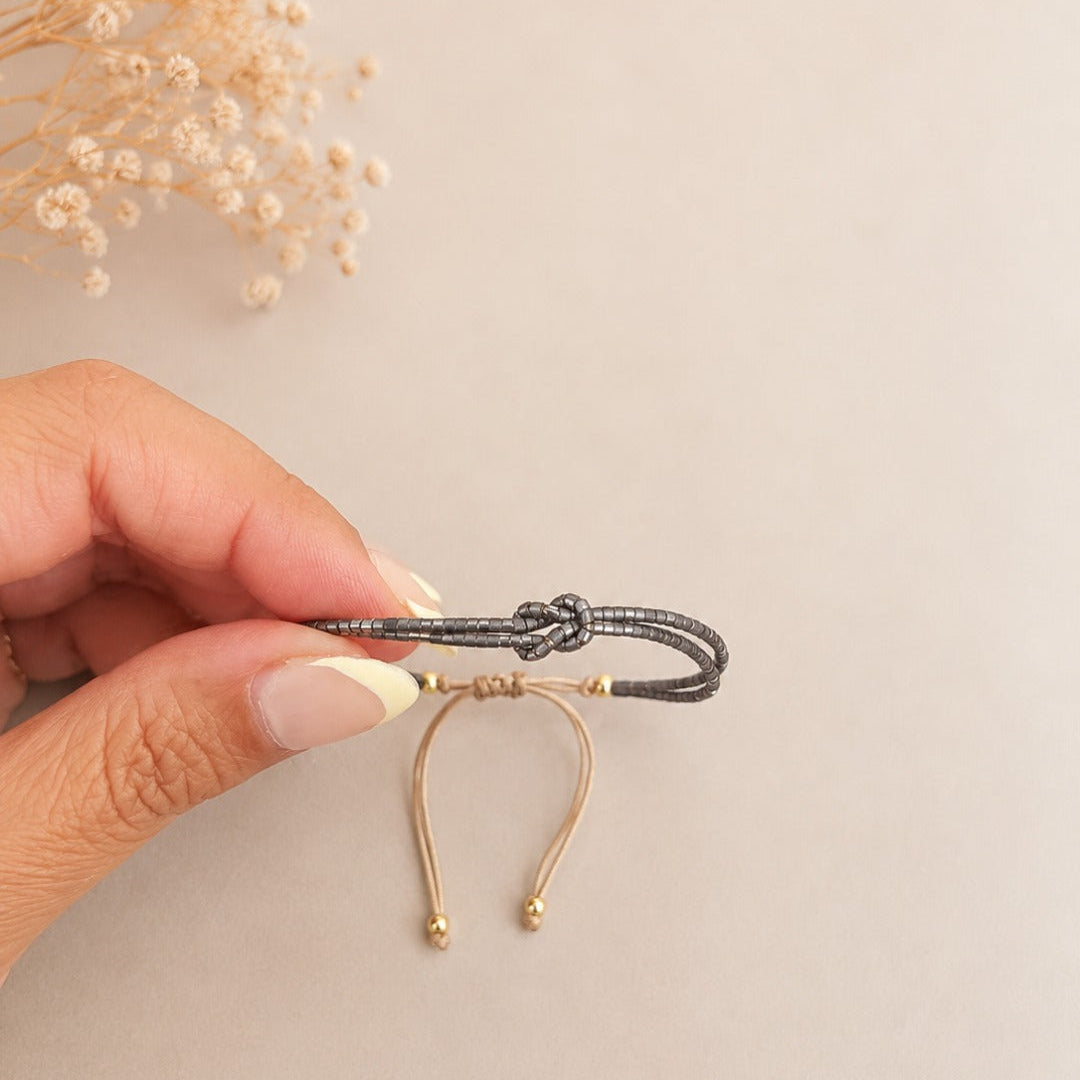 A hand holding a black braided cord bracelet with a knot closure and small gold bead accents, shown against a light beige background with dried flowers in the corner.