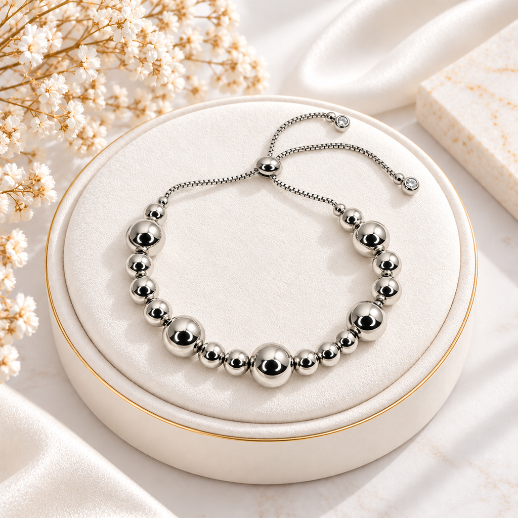 A silver beaded bracelet with a matching adjustable chain and slider clasp, displayed on a round cream jewelry cushion with dried white flowers and satin fabric in the background.