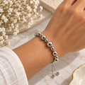 A polished silver beaded bracelet with a sliding chain and bead toggle, worn on a person's wrist. The background features a soft-focus arrangement of white baby's breath flowers and a light-colored textured surface.
