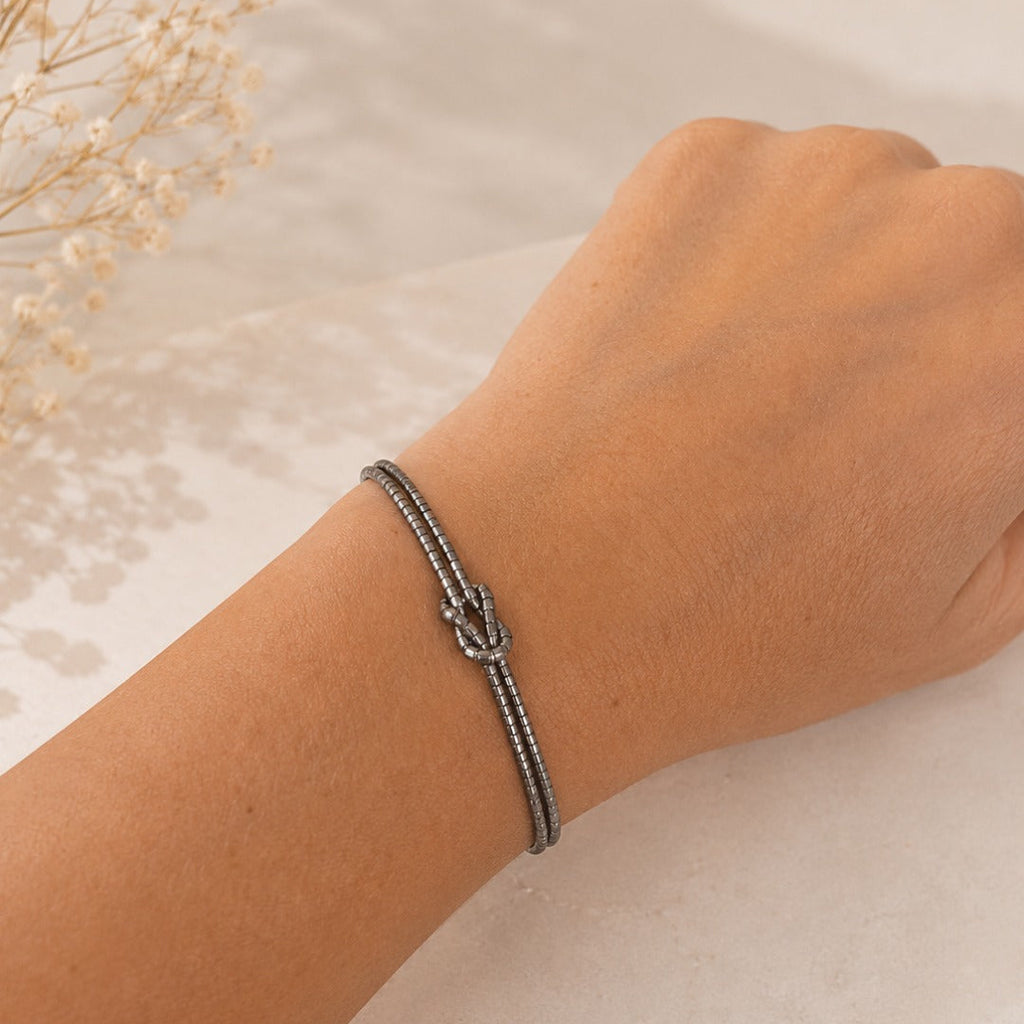 A close-up of a person's wrist wearing a silver-toned rope bracelet with a knotted center detail, resting on a light marble surface with dried flowers in the background.