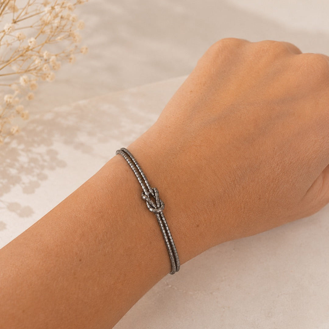 A close-up of a person's wrist wearing a silver-toned rope bracelet with a knotted center detail, resting on a light marble surface with dried flowers in the background.