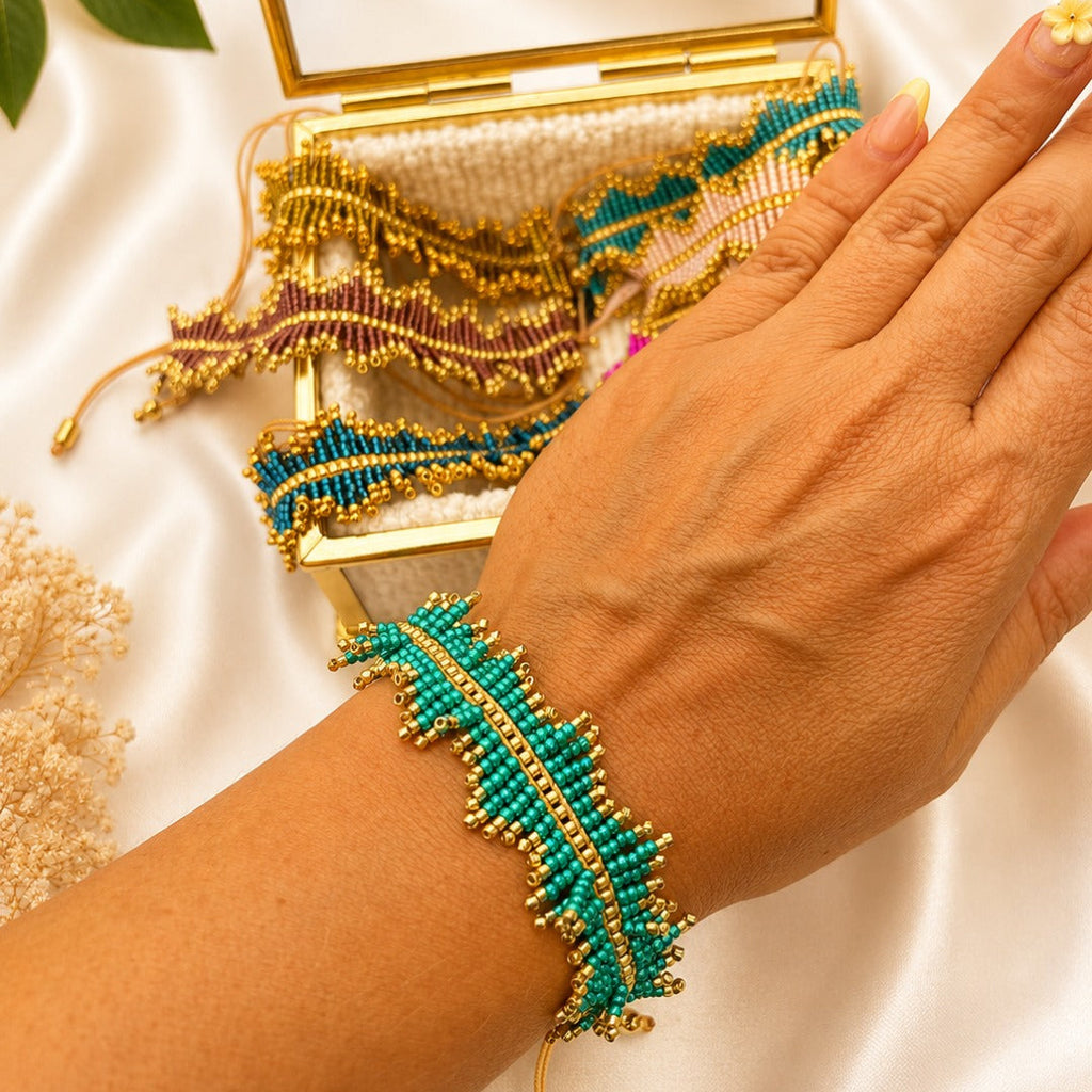 A hand wearing a turquoise and gold beaded leaf-shaped bracelet, with a matching open jewelry box and additional bracelets on a white satin background.